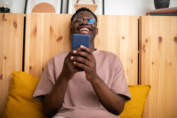 Young black man laughing at home relaxing using mobile phone to watch funny videos. Reading text...