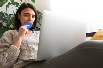Pensive young woman in doubt holds credit card wondering if buying online.Checking online bank...