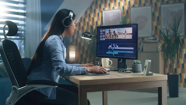 Asian woman in headphones editing video for blog or documentary or feature film about space in professional video editing software on computer, while sitting at the table at home and working remotely.