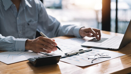 Close-up of a businessman sitting at a desk concentrating on fluently inspecting work using his laptop and several documents on the table at the office