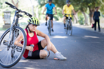 Exhausted old woman road cyclist in protective sportswear sitting on the ground and drinking water...