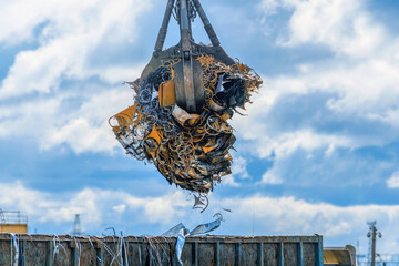 A grab crane loads iron, steel and scrap metal into a truck for recycling at an industrial waste collection center with small falling particles of metal chips and dust. Transportation of metal waste. - obrazy, fototapety, plakaty