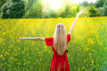 Beautiful girl in a flowering field in a red dress with arms wide open. Sunbeams, glare, light leaks and sun rays. Young woman feeling free in the field with flowers in sunshine, freedom peace