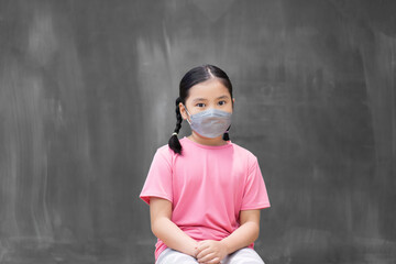 Asian child student or kid girl wearing dark face mask for protect dust or covid-19 to sitting on front dirty chalkboard or empty old blackboard for teach study in classroom at school with copyspace