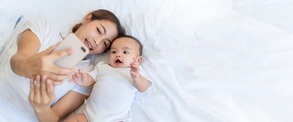 Authentic close up of asian mother and her newborn baby making a selfie or video call to father or relatives in a bed.Concept of technology,parenthood,family,connection,technology,Video Call.
