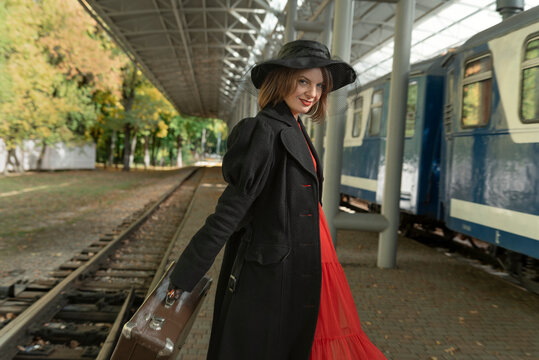 Young woman in red dress, black retro coat and hat holds vintage suitcase and stands on platform near train.