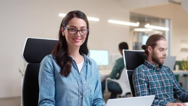 Camera approaching young Caucasian beautiful happy woman in glasses typing on laptop looking at camera and smiling. Professional people work in modern office. Business. Close up. Worker concept