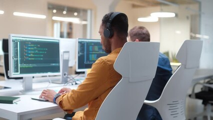 Rear of young man IT programer working on desktop computers typing on keyboard coding a program and listening to music in headphones. Close up. Professional worker writing website in modern office