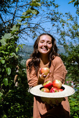 Happy young healthy woman holding straw hat full of fresh organic apples in sunny day in garden. Female wellness and autumn harvest concept.