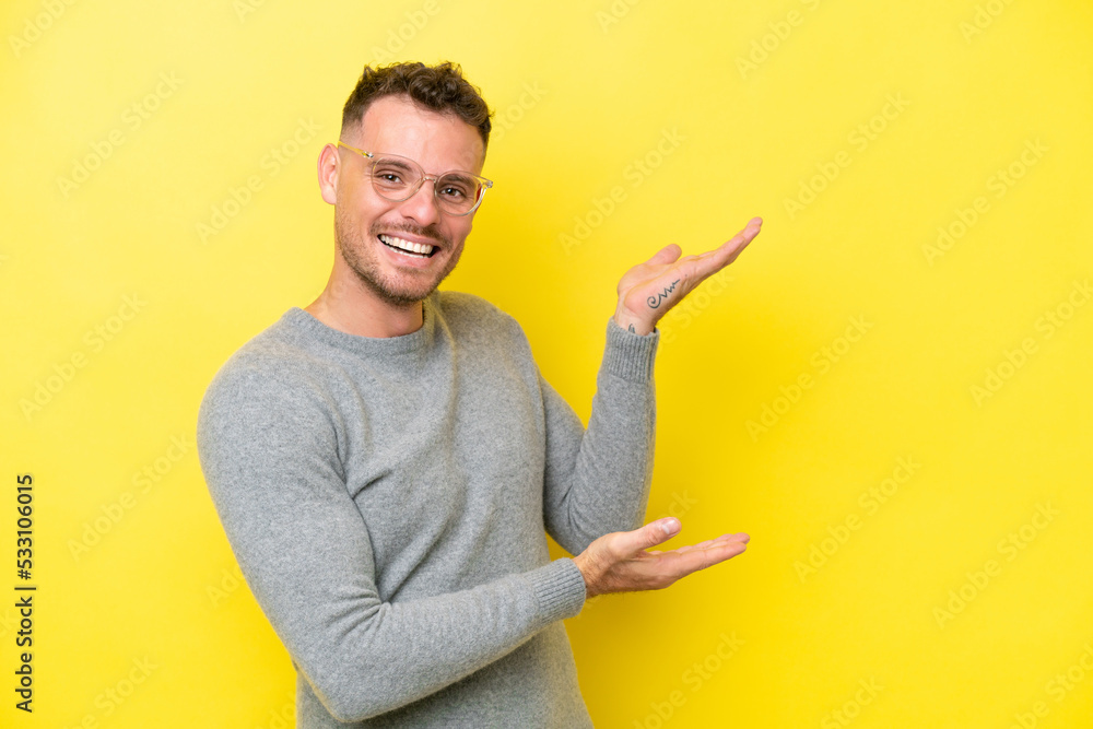 Wall mural young caucasian handsome man isolated on yellow background extending hands to the side for inviting 