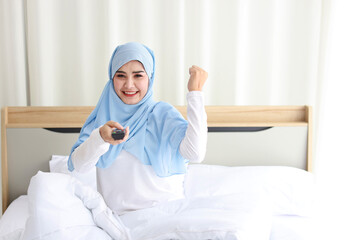 TV and happiness concept. Beautiful asian muslim woman in white sleepwear sitting on bed, holding television remote and raise hand, shout loudly and celebrate something with glad happy smiling face.