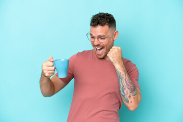 Young caucasian man holding cup of coffee isolated on blue background celebrating a victory