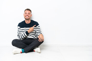 Young man sitting on the floor isolated on white background pointing to the side to present a product