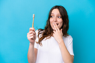 Young caucasian woman brushing teeth isolated on blue background with surprise and shocked facial...