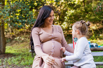 Happy pregnant mother and daughter hugging on a park bench. Happy motherhood. Beautiful woman with a little daughter in nature