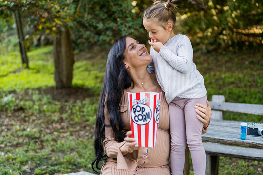 A Pregnant Woman And Her Daughter Are Sitting On A Bench In An Autumn Park And Eating Popcorn, Having Fun Together.