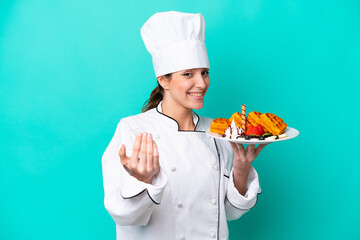 Young caucasian chef woman holding waffles isolated on blue background inviting to come with hand. Happy that you came