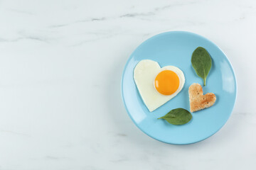 Romantic breakfast with heart shaped fried egg and toast on white marble table, top view. Space for text