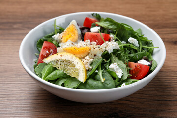 Delicious salad with boiled egg, tomatoes and cheese in bowl on wooden table, closeup