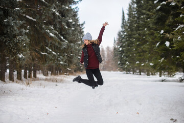 happy jumping girl in a blue hat, sweater and vest, on a snowy winter alley among the fir trees