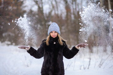 blonde girl in a black fur coat and a blue hat against the backdrop of a winter snowy forest. She...