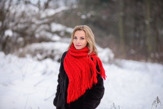 blonde girl in a black fur coat and a red scarf against the backdrop of a winter snowy forest