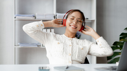 Asian woman wearing headphones to listen to music in her break from work, she is in the office of...