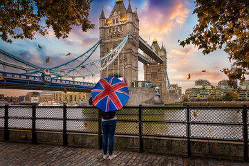 London in autumn time concept with a tourist person holding a british flag umbrella in front of the famous Tower Bridge during sunset time