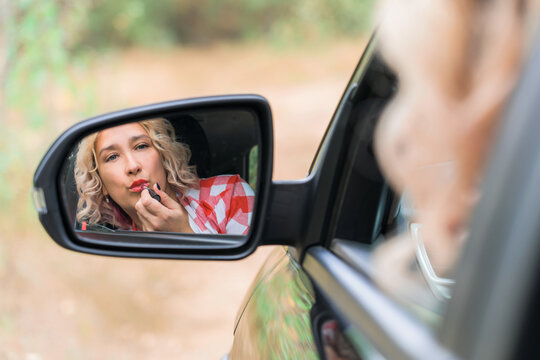 A woman sitting behind the wheel of a car paints her lips looking in the side mirror