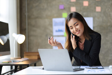 Attractive Asian businesswoman talking on mobile phone working  in modern office. Happy business woman talking on mobile phone while analyzing monthly work schedule in laptop.