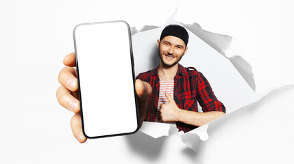 Studio portrait of young smiling man, showing big smartphone with blank on screen through torn...