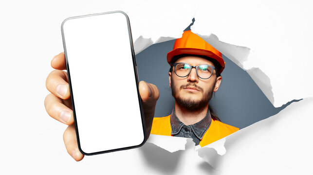 Studio portrait of young man, construction worker, showing big smartphone with blank on screen through torn paper hole. White background. Wearing eyeglasses and safety equipment.