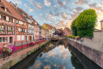Atardecer en la Petite Venise de Colmar, Alsacia, con las calles llenas de flores, turistas paseando y un bonito cielo con nubes reflejadas en el agua del canal