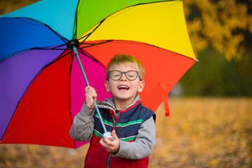 child boy in glasses, standing under a rainbow umbrella, against the background of autumn leaves