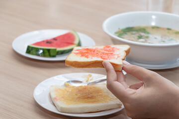 Hand holding slice of toast with strawberry jam, served with honey toast, watermelon, and hot soup on wooden table, healthy homemade breakfast concept, traditional Asian morning meal