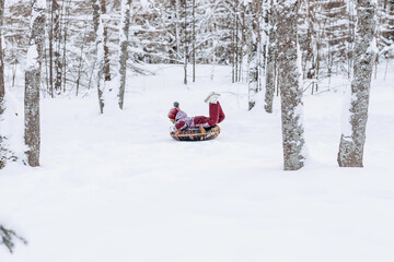 Happy little African-American girl in a red hat and jumpsuit rides on tubing in the winter park.Beautiful trees are covered with white snow.Winter fun concept.Selective focus,copy space.