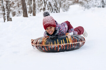 Happy little African-American girl in a red hat and jumpsuit rides on tubing in the winter...
