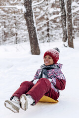 Fototapeta na wymiar Happy little African-American girl in a red hat and jumpsuit rides a saucer in the winter park.Beautiful trees are covered with white snow.Winter fun,active lifestyle concept.