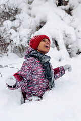 Fototapeta na wymiar Happy little African-American girl in a red hat and jumpsuit walks in the winter forest.Beautiful trees are covered with white snow.Winter fun,active lifestyle concept.