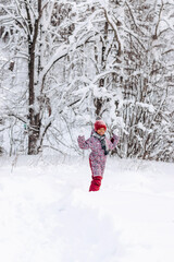 Fototapeta na wymiar Happy little African-American girl in a red hat and jumpsuit walks in the winter forest.Beautiful trees are covered with white snow.Winter fun,active lifestyle concept.Selective focus,copy space.