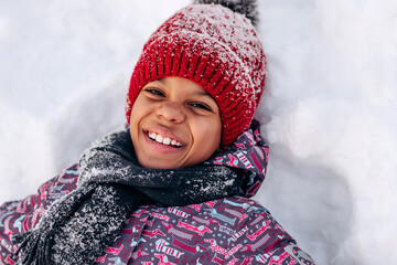 Happy little African-American girl in a red hat and jumpsuit is lying in the snow.Winter fun,active...