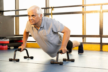 Fototapeta na wymiar Active old man in sportswear doing push ups with bars at gym 