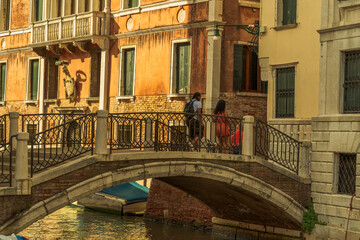 Tourists on the bridge over the canal street in Venice, old walls of houses with windows on the Venetian street in the morning sunlight, stone bridge over the canal of the city of Venice
