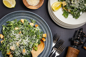 Kale Cesar salad served as a family style in a bowl with stack of white plates on the side. Black pepper grinder, lemon slice and grated Parmesan as a props.
