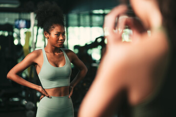 Young black lady wearing sportswear looking at the mirror in the gym with anxious expression.