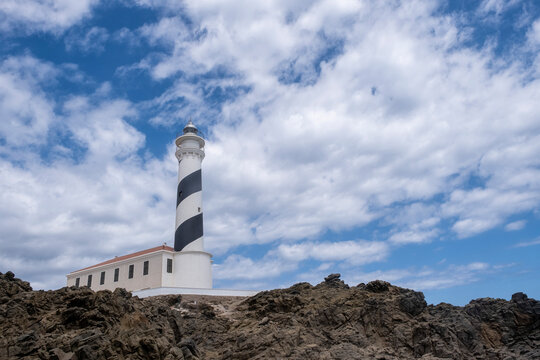 Favaritx lighthouse with its tower painted with a blue and white spiral on a rocky cliff, with the sky of cotton clouds, low point of view, copy space, horizontal, Menorca, Spain