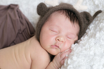 A cute little baby in a knitted hat with deer horns sleeps on a white bouclé bedspread at home. Health and motherhood