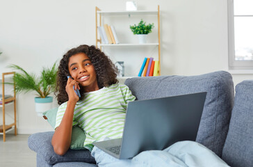 Fototapeta premium Dark-skinned teenage girl is having fun talking on phone and using laptop at same time. African American girl uses modern wireless technology while sitting on sofa at home in living room.