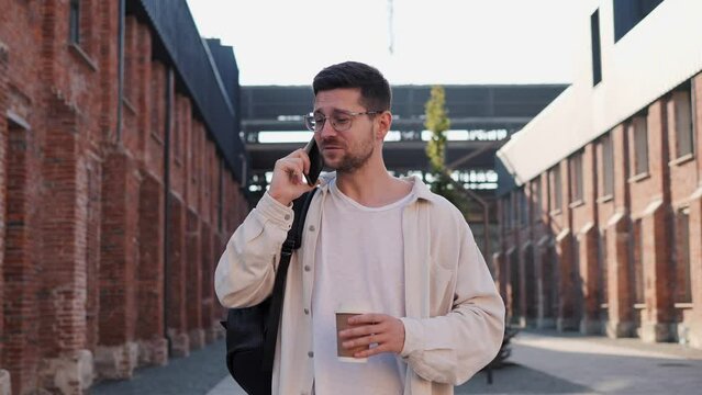 Portrait of attractive man in casual clothes in city talking on smart phone outdoors. Man in glasses holding hot drink to go and talking on smartphone while walking on steps on city street. 