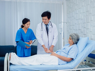 Obraz premium Asian male doctor, professional in white lab coat holding elderly patient's hand for check to examine heartbeat pulse and nursing assistant taking note condition in hospital. Health follow-up concept.